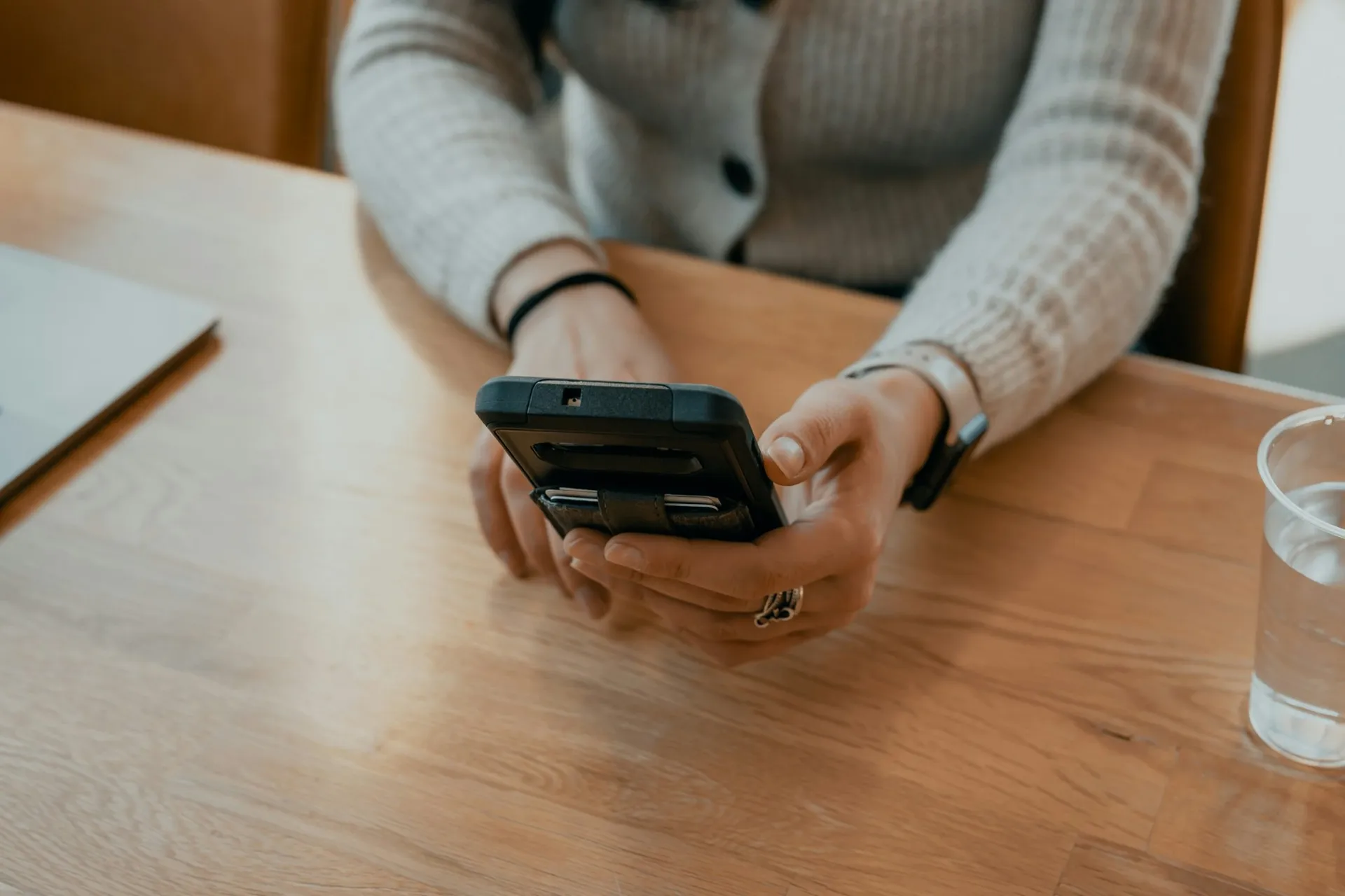 Hands holding a phone on a wooden table beside a laptop and a glass of water